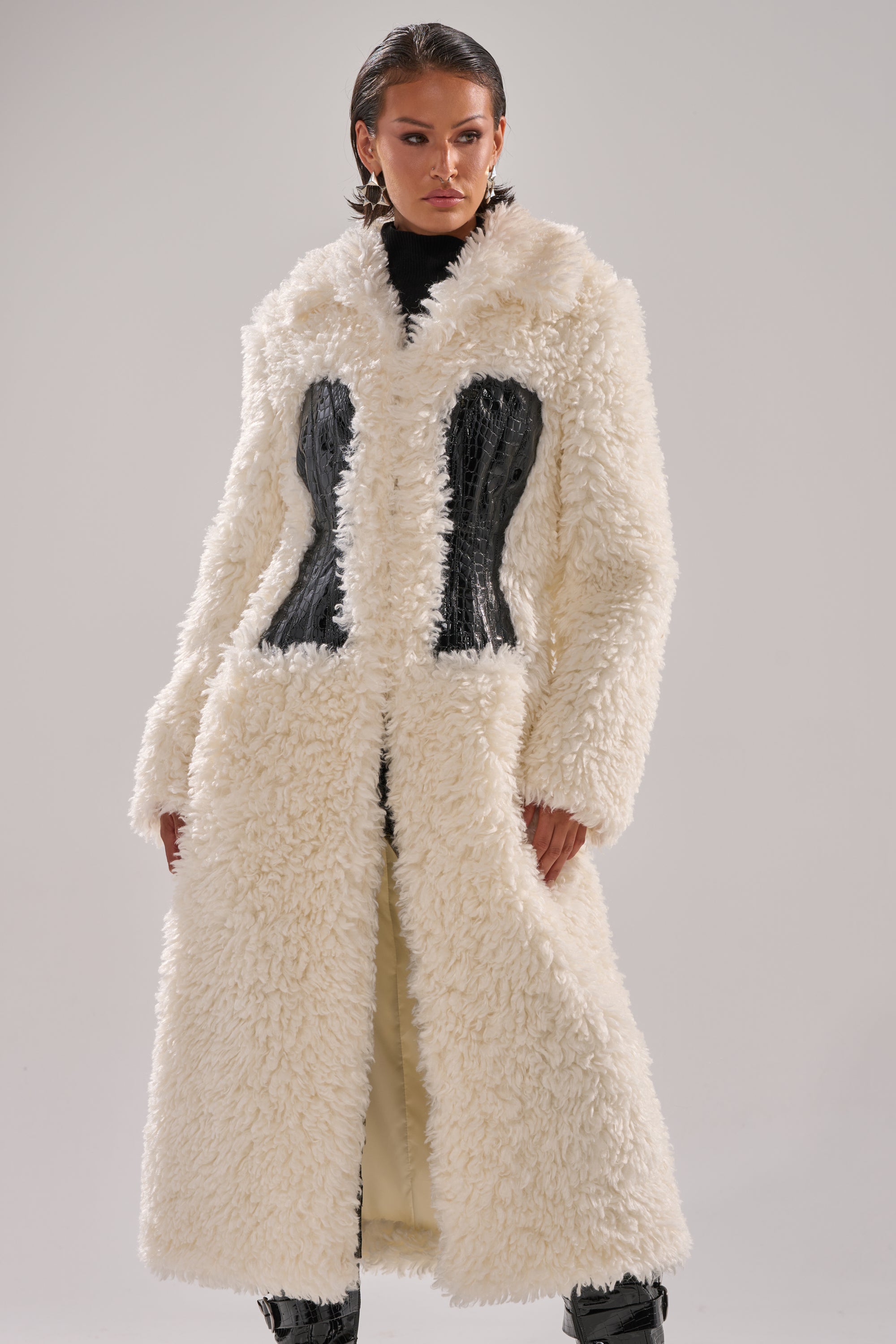 A woman wears the BUY ME ROSES CORSETED FUR COAT—long, white, and fluffy with black crocodile leather paneling—over a black turtleneck, standing against a plain light background.