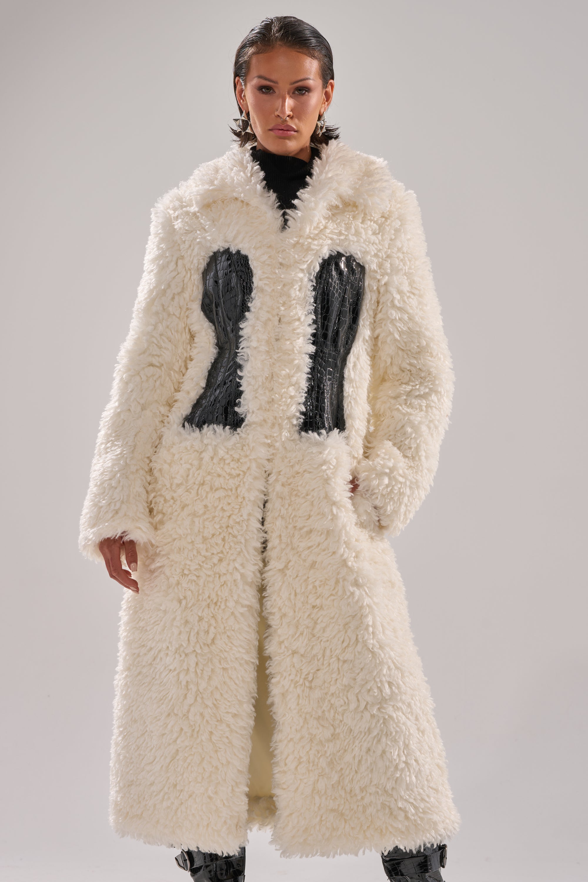 A person models the BUY ME ROSES CORSETED FUR COAT, a long white faux fur coat with black crocodile leather panels, against a plain light gray background.