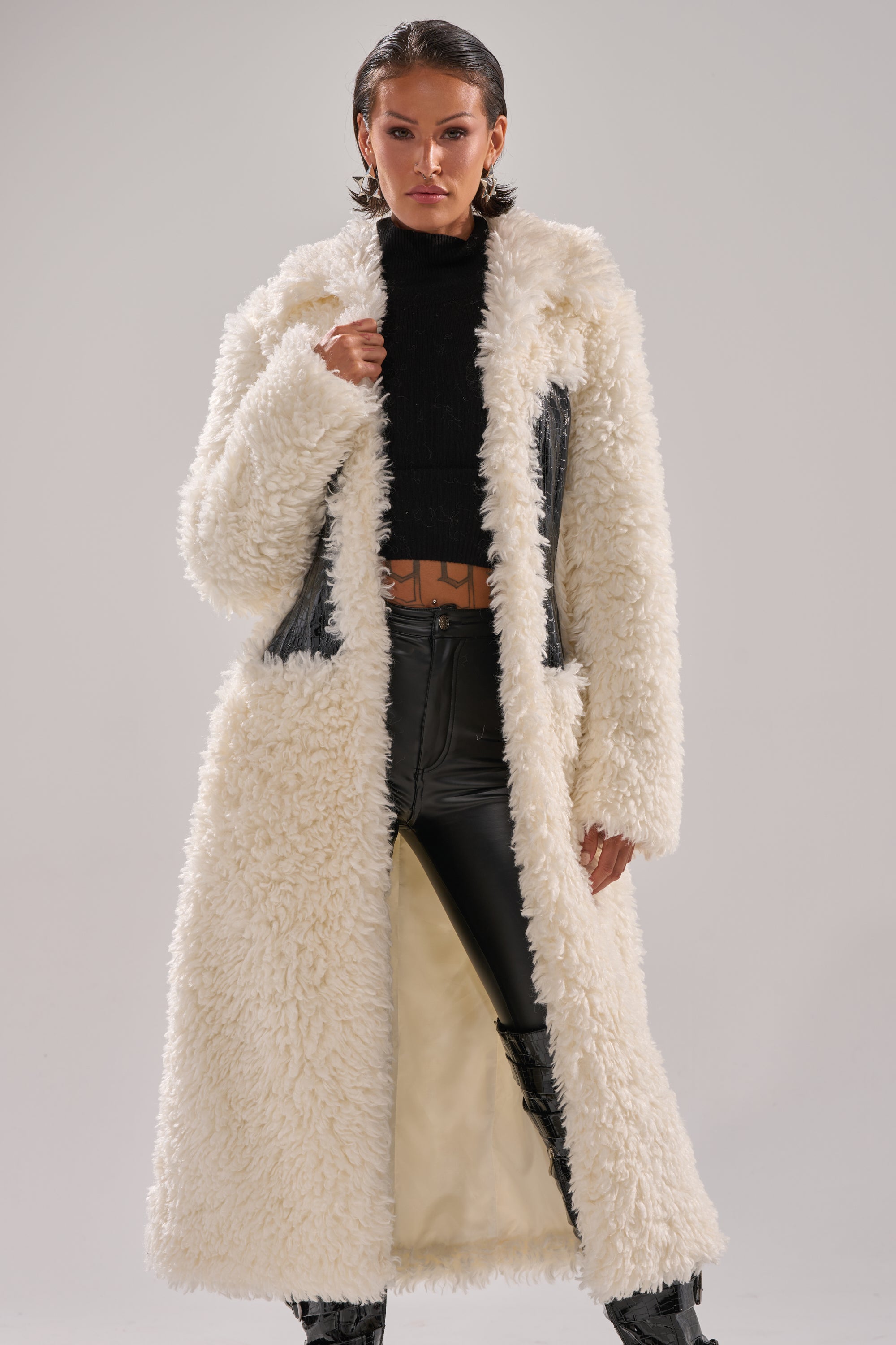 A person wears the BUY ME ROSES CORSETED FUR COAT over a black cropped top and high-waisted black leather pants with subtle crocodile paneling, standing confidently against a plain background.