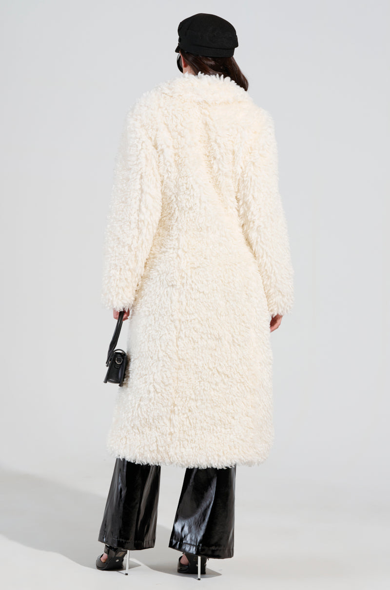 Dressed in the BUY ME ROSES CORSETED FUR COAT, a person stands facing away in their ensemble of black pants. They complement their look with a black cap and hold a small black handbag, all set against a plain white background that highlights the sleek style of their outfit.