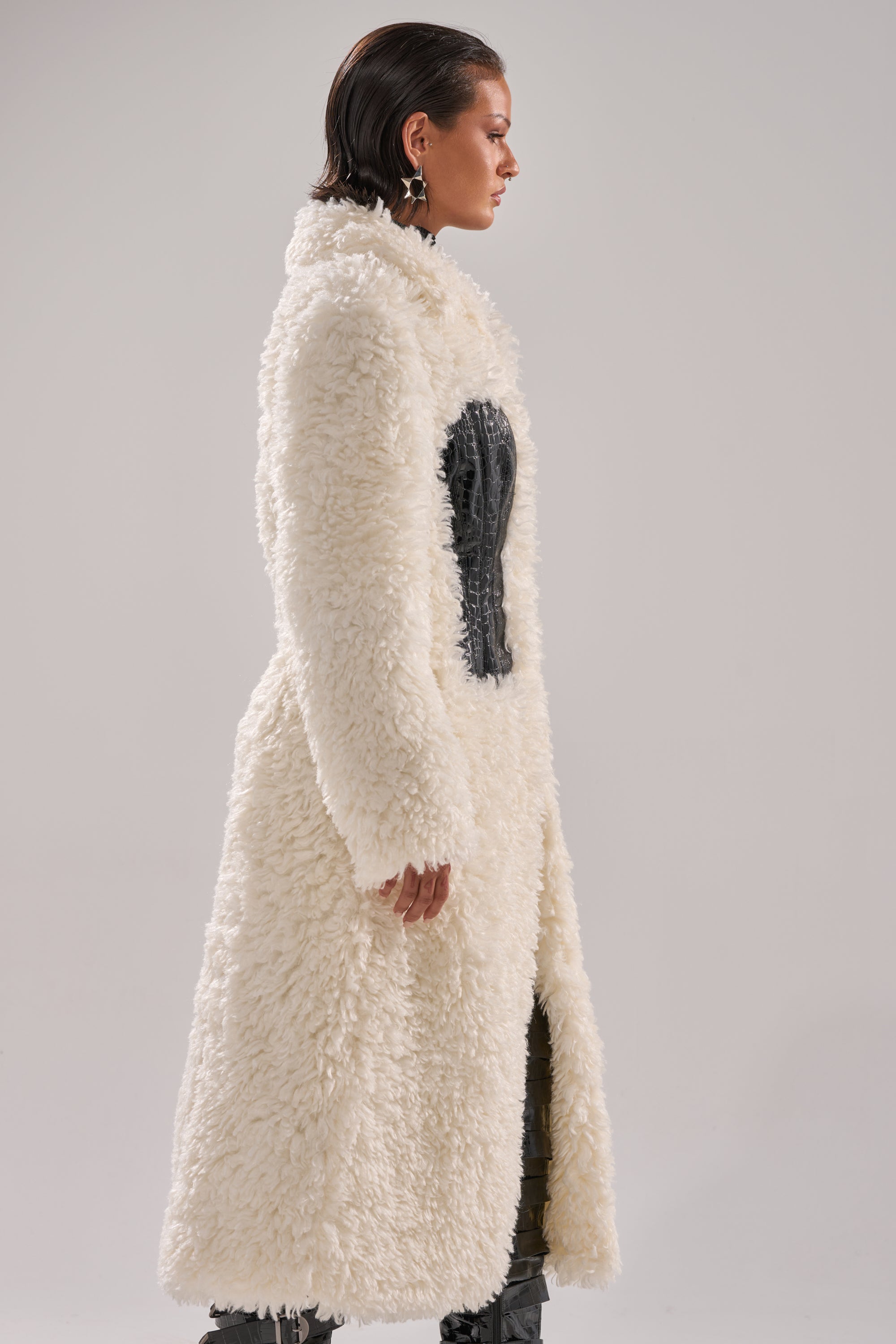 A person stands in profile wearing the BUY ME ROSES CORSETED FUR COAT—long, textured white faux fur with black details and crocodile leather chest paneling—paired with dark pants and statement earrings against a plain light background.