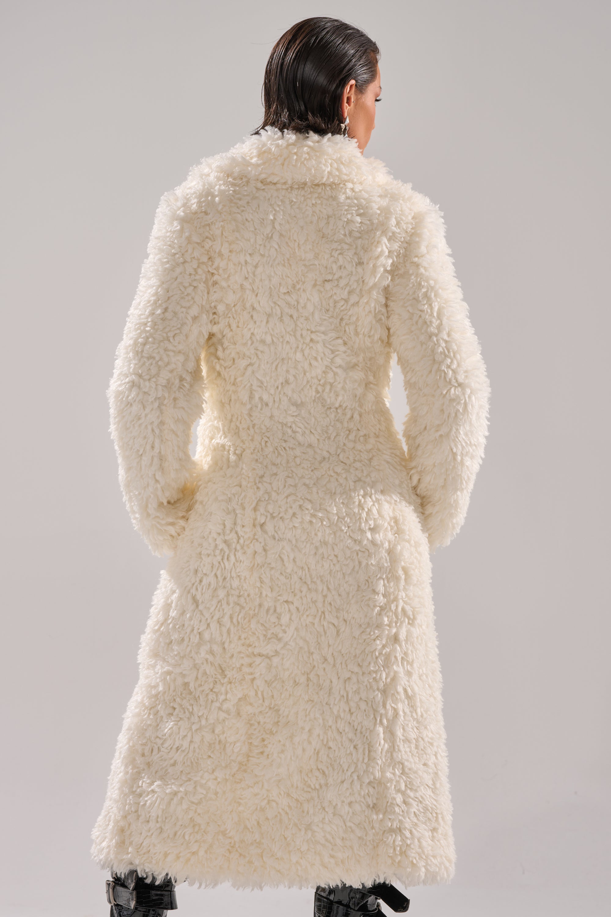 A person with wet, slicked-back hair is seen from behind in the BUY ME ROSES CORSETED FUR COAT—an off-white faux fur piece—paired with black boots, set against a light gray studio background.