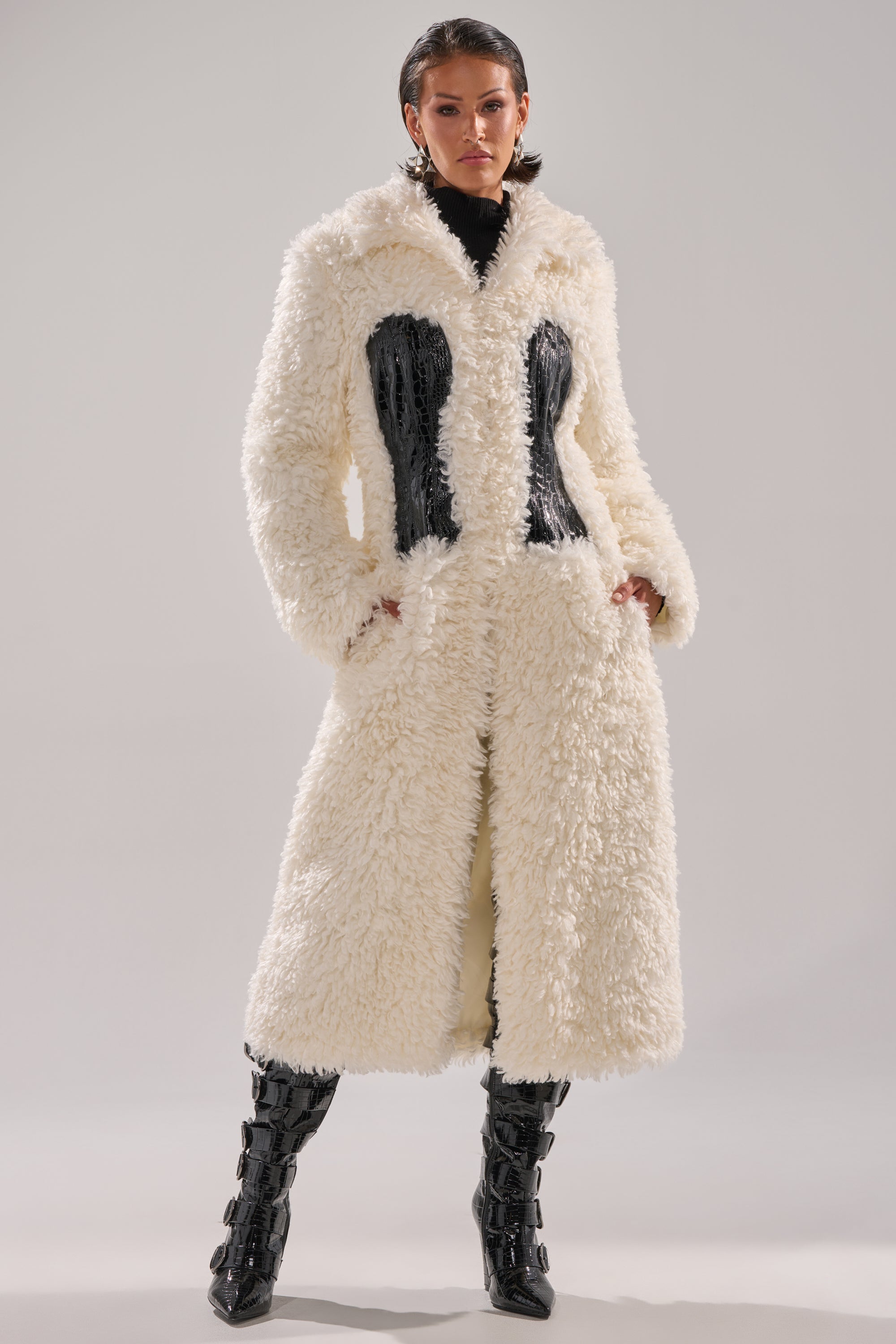 Someone stands confidently in the BUY ME ROSES CORSETED FUR COAT—a white, fluffy faux fur piece with built-in corset detail—paired with black patent knee-high boots against a plain, light background.