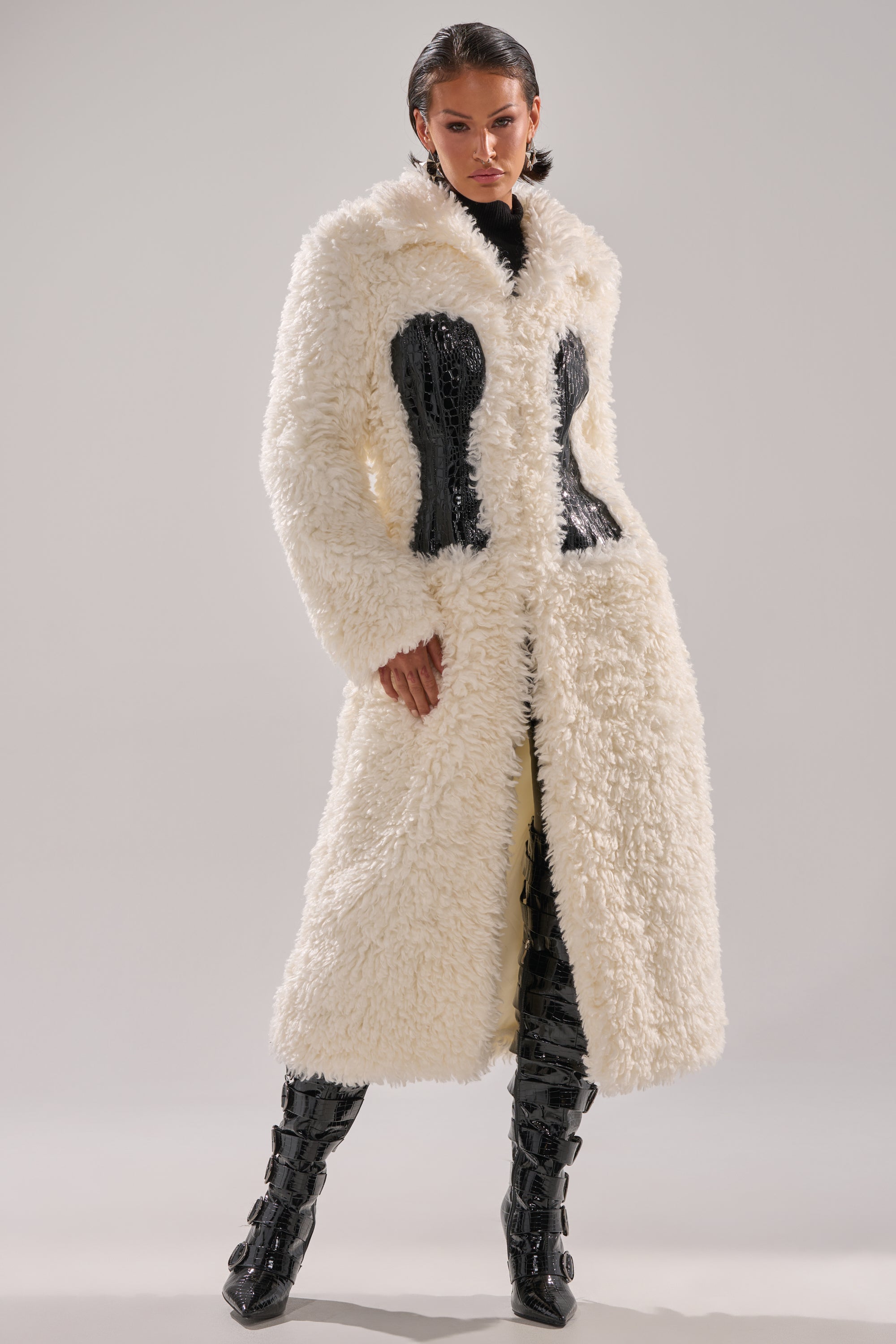 A woman poses in the BUY ME ROSES CORSETED FUR COAT, a long white fur style with black corset detailing, paired with black textured boots. Her hair is slicked back as she stands against a plain light background.