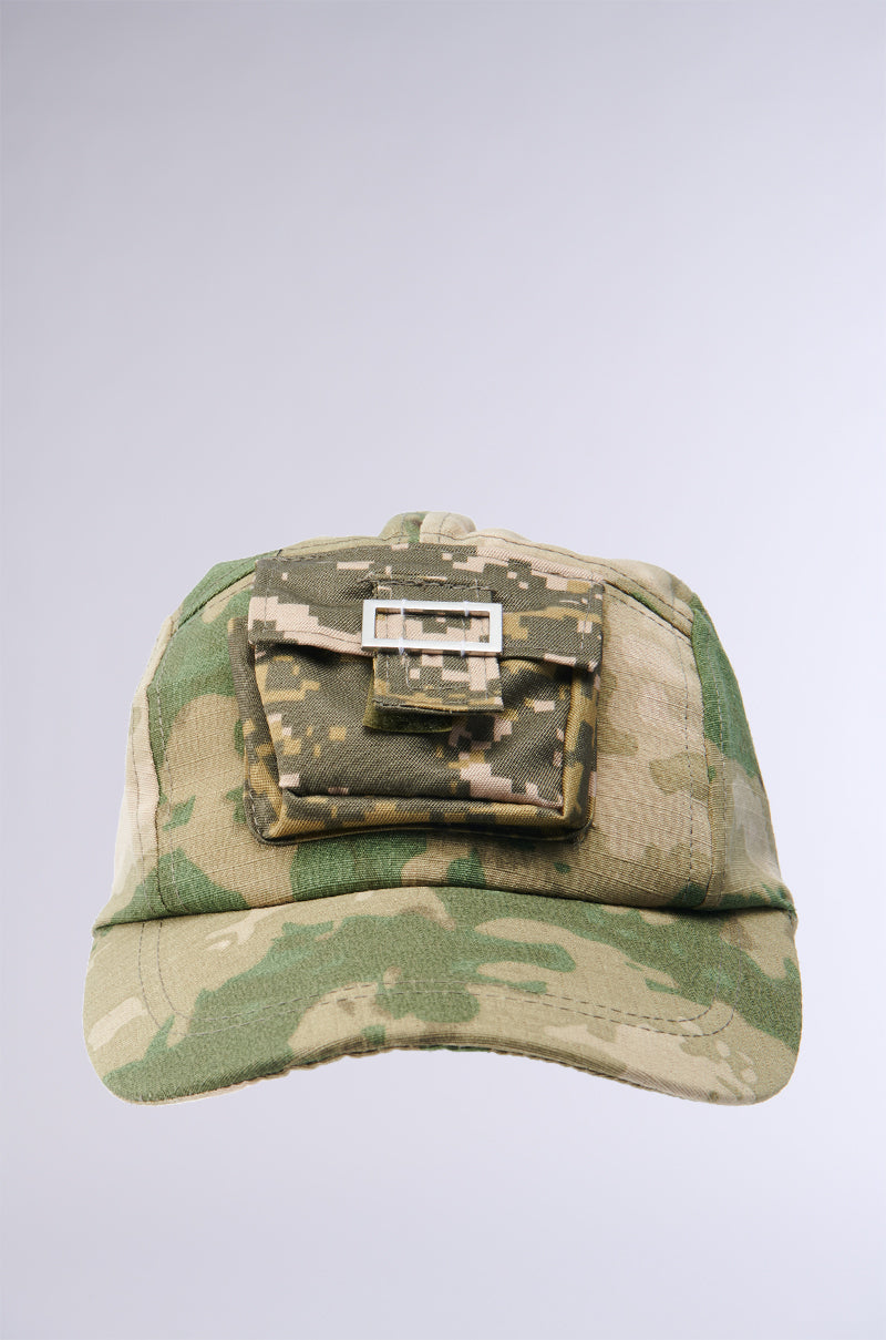 The TRY AND FIND ME CAMO SNAPBACK showcases a pixelated camouflage design in green and beige, complete with a small rectangular pocket on the front that features a flap secured by a buckle. It offers an adjustable Velcro strap for the perfect fit, all set against a plain light gray background.