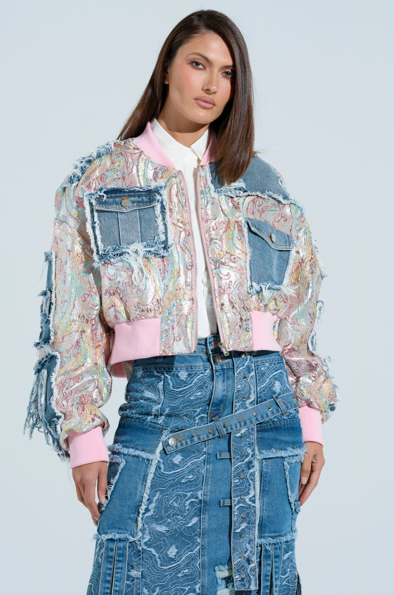 A woman models a vibrant SPRING REFRESH BROCADE BOMBER with pink cuffs and denim patches over a patchwork denim skirt. She has straight, shoulder-length hair and stands against a plain backdrop.