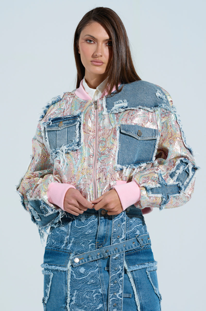 Wearing the SPRING REFRESH BROCADE BOMBER, a woman with long hair dons a colorful patchwork jacket featuring denim accents, metallic-infused textured fabric, and raw edges. She pairs it with a matching skirt against a light blue background, embodying a pastel daydream while looking directly at the camera.