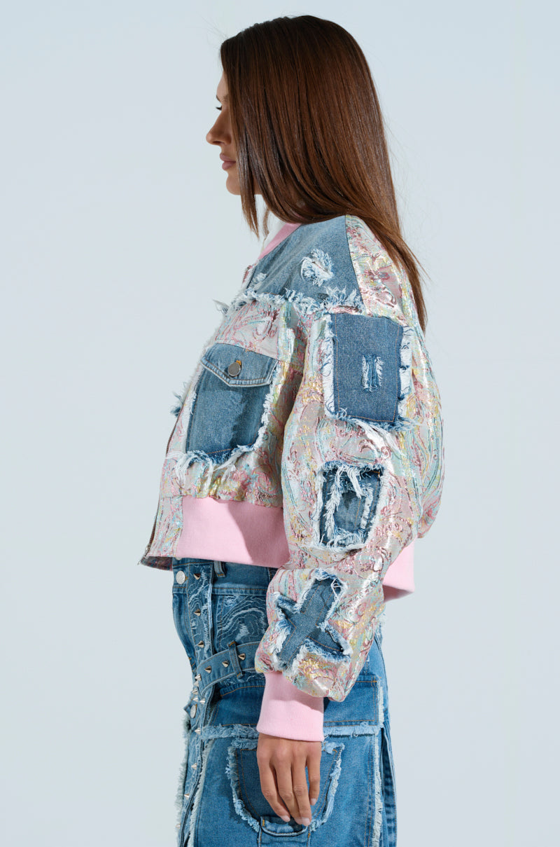 A person stands in profile, wearing the SPRING REFRESH BROCADE BOMBER jacket with a colorful pastel daydream vibe and pink trim, paired with a matching patchwork denim skirt, against a plain background.