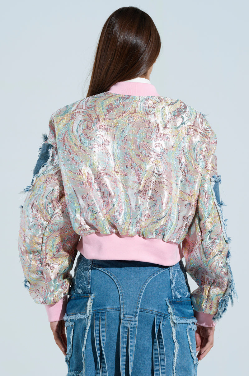 A person with long hair wears the SPRING REFRESH BROCADE BOMBER, a colorful metallic-infused jacket with paisley patterns and pink accents. They pair it with uniquely designed, frayed blue jeans. Captured from behind against a light backdrop, the image exudes a Pastel Daydream vibe.