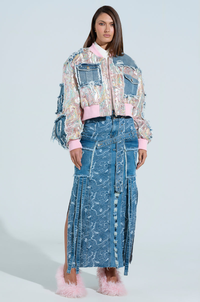Against a light blue background, a woman wears the SPRING REFRESH BROCADE BOMBER with a long, patterned denim skirt and pink fluffy sandals. Her neutral expression complements her straight dark hair.