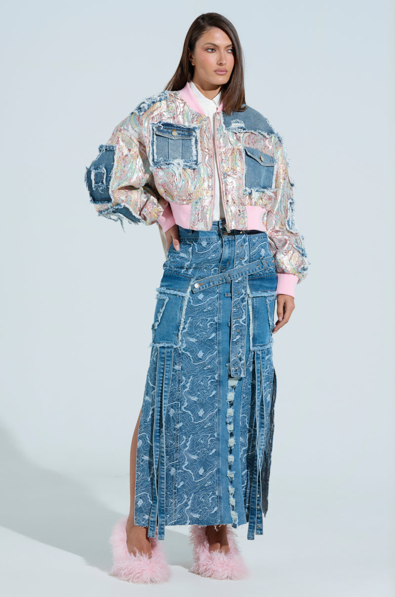 A woman stands wearing the SPRING REFRESH BROCADE BOMBER, a textured, multicolored cropped jacket, and a long patchwork denim skirt with a front slit. Her metallic-infused pink fuzzy slippers add flair, while the light pastel blue background enhances the dreamy scene.