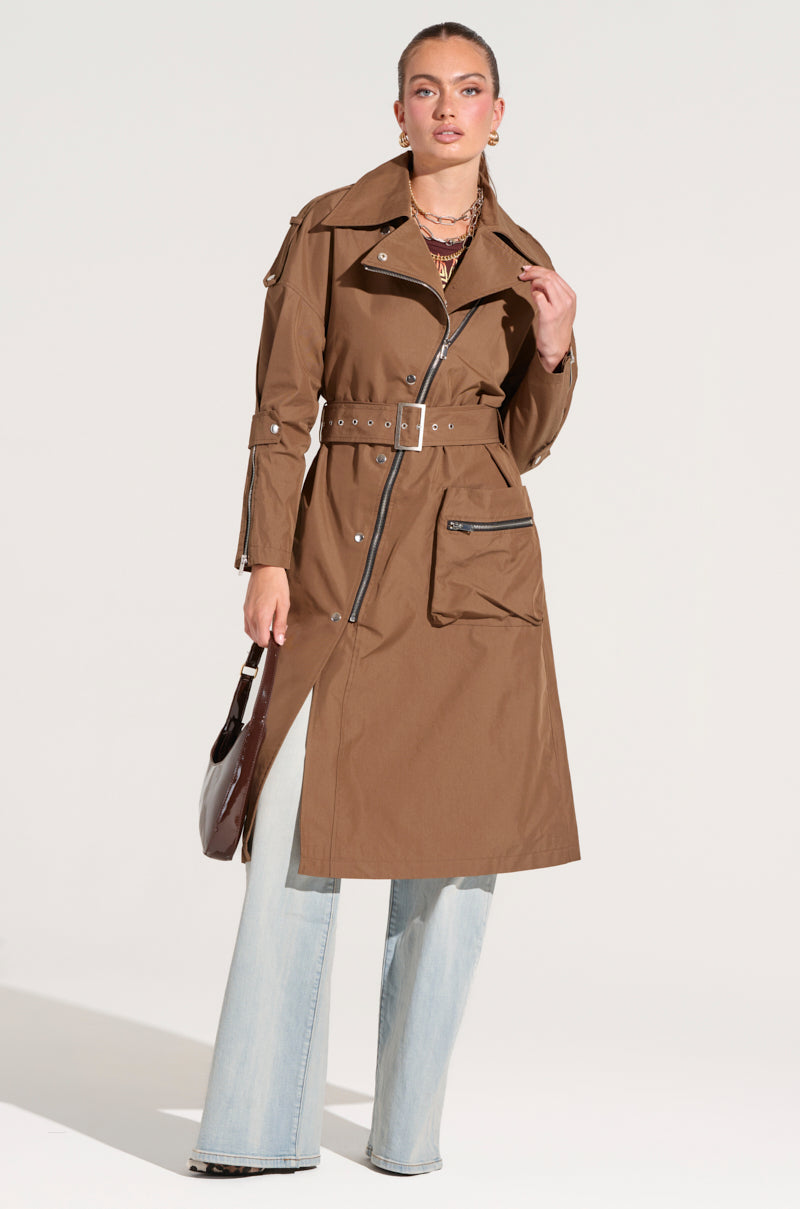 A woman stands confidently wearing the stylish MANHATTAN ASYMMETRICAL TRENCH coat, which features large pockets. Paired with light-washed flared jeans and holding a dark handbag, her look exudes city chic outerwear vibes. Crafted from a cotton blend fabrication, she poses elegantly against a plain light-gray background with her hair pulled back.