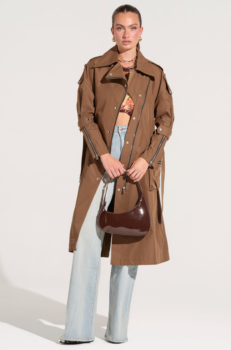 A woman is wearing a long brown MANHATTAN ASYMMETRICAL TRENCH over a floral crop top and light blue jeans. She is holding a dark brown handbag and has her hair pulled back, exuding city chic outerwear. She stands against a plain white background.