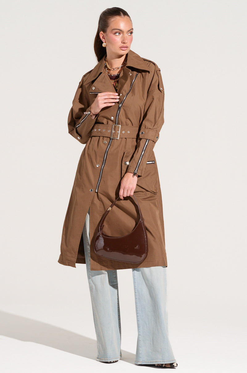 A woman stands confidently wearing the MANHATTAN ASYMMETRICAL TRENCH with a belt, paired with light blue wide-leg jeans and black shoes. She carries a glossy dark brown handbag and her hair is pulled back. The plain, light background highlights her city chic outfit perfectly.