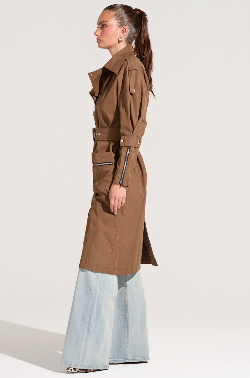 A person with a ponytail stands in profile wearing the MANHATTAN ASYMMETRICAL TRENCH with belted cuffs and pockets, over light blue flared jeans, and leopard print shoes. They are facing to the left against a plain white background.