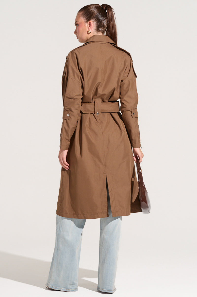 A person with a high ponytail is wearing the Manhattan Asymmetrical Trench, epitomizing city chic style. The long brown trench coat, distinguished by its belt and wide sleeves, is paired with light blue jeans. They're holding a brown handbag while standing and facing away from the camera against a plain background.