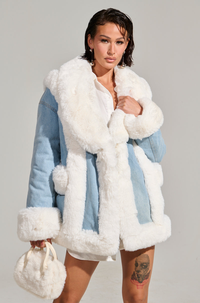 A person stands confidently wearing the SITTING PRETTY FUR TRIM COAT, a light blue creation with white fur trim, over a white shirt. They carry a small, fluffy bag and have a tattoo on their left thigh. The background is a plain, neutral color.