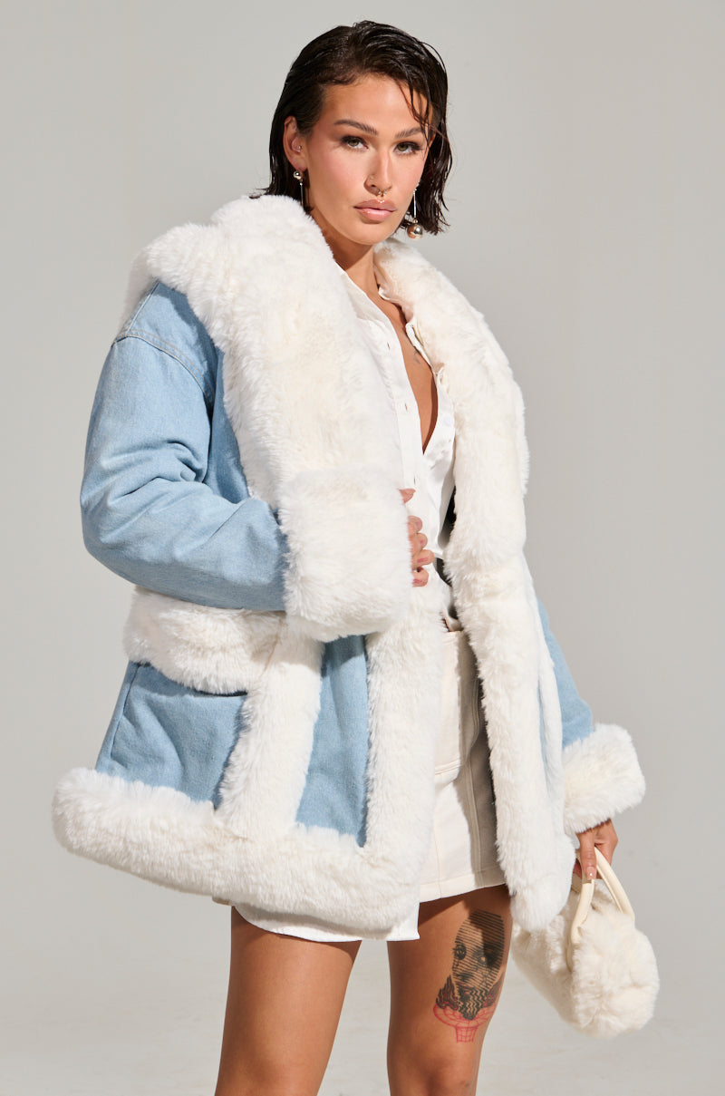 A person wearing a fashionable light blue SITTING PRETTY FUR TRIM COAT with fluffy white fur trim over a white outfit holds a small matching fur-accented bag. They have short dark hair, one leg adorned with a colorful tattoo, and are standing against a plain background.