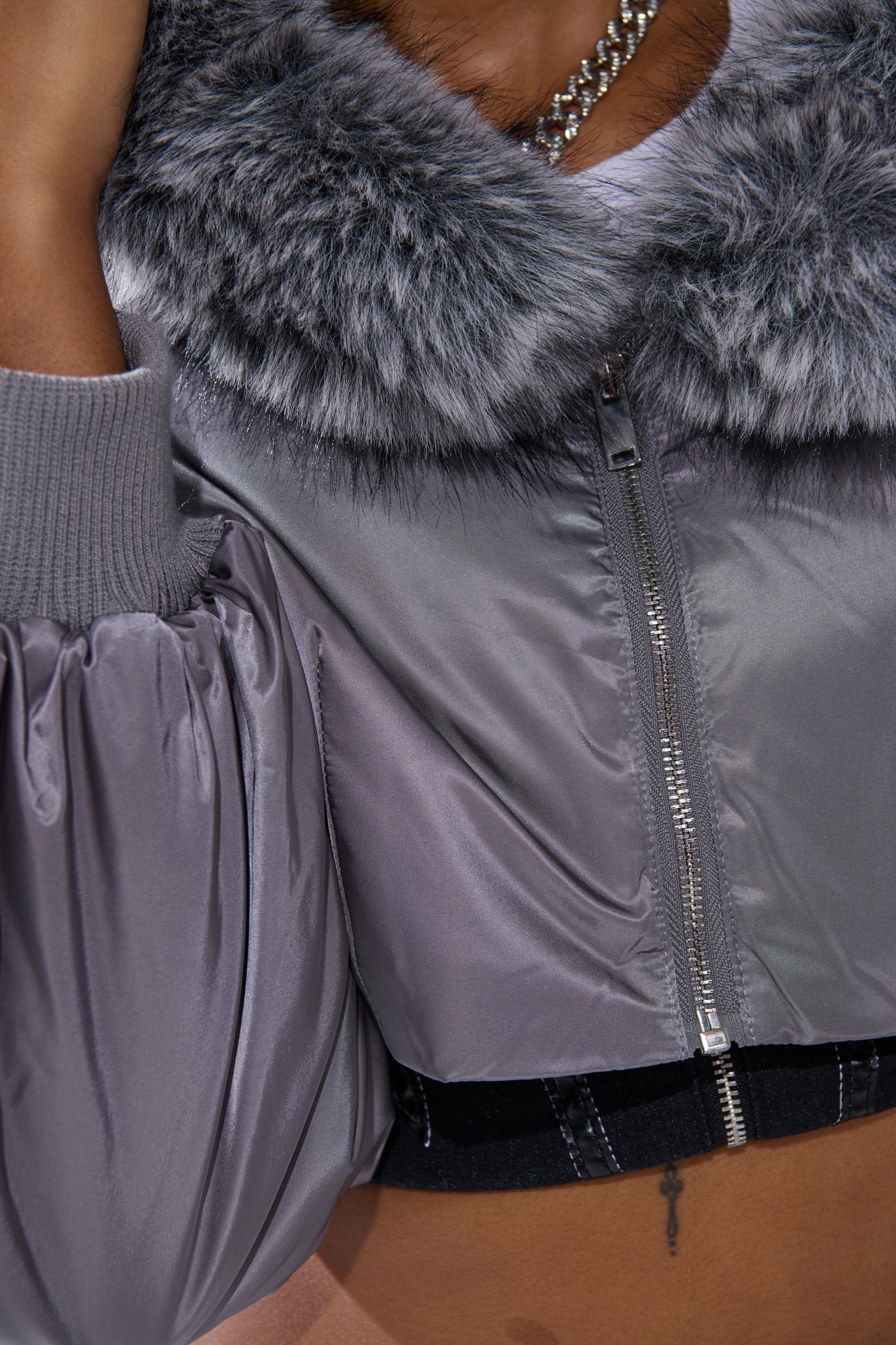 Close-up of a person rocking the ITTY BITTY CROPPED BOMBER in shiny silver, featuring a luxe fur-trim collar, ribbed cuffs, and a zipper. Styled with a silver chain necklace and bare midriff for an edgy street-style vibe.