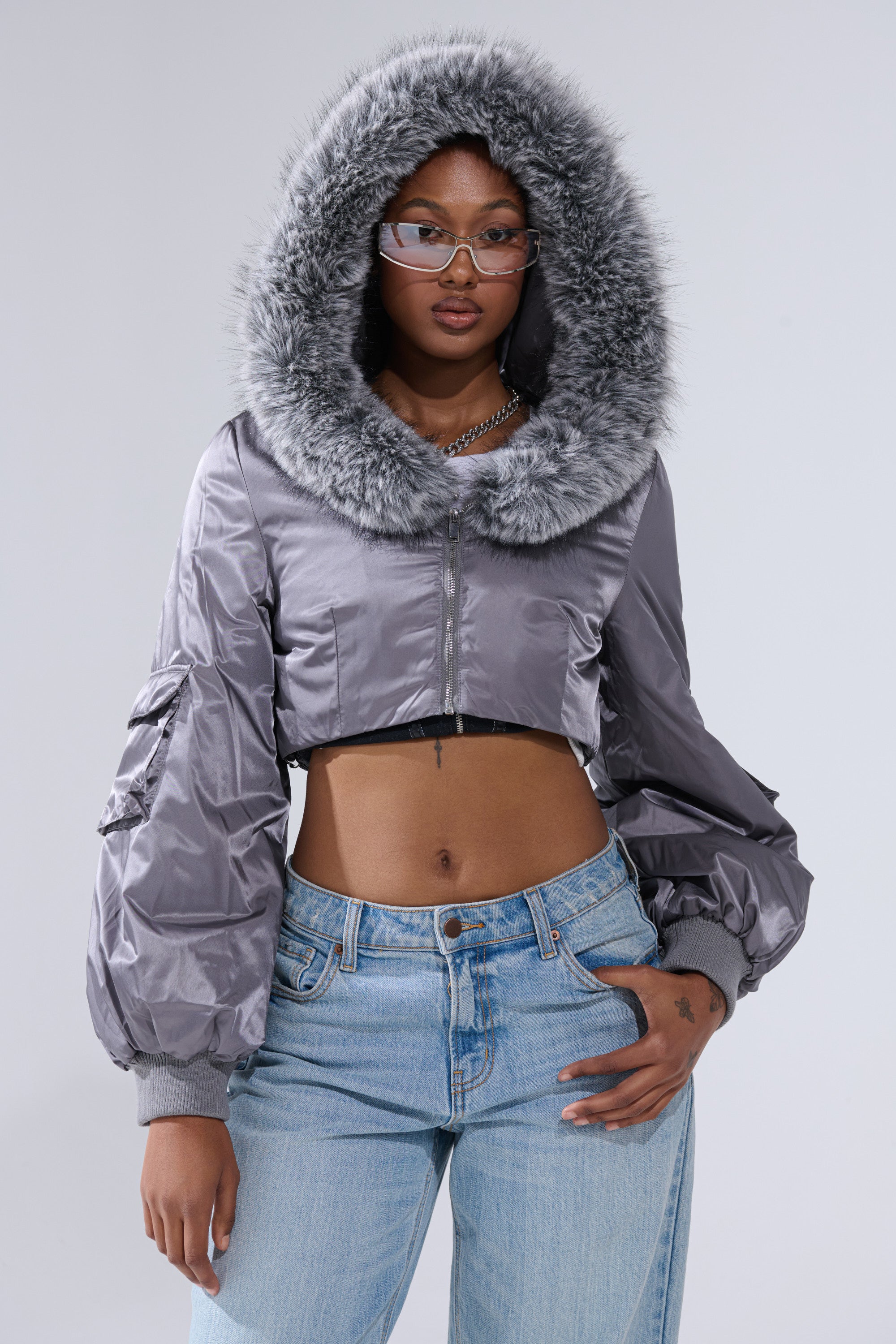 A woman wears the ITTY BITTY CROPPED BOMBER, a shiny gray bomber jacket with fur trim, styled with blue jeans, clear glasses, and a chain necklace against a light gray background for a chic street style look.