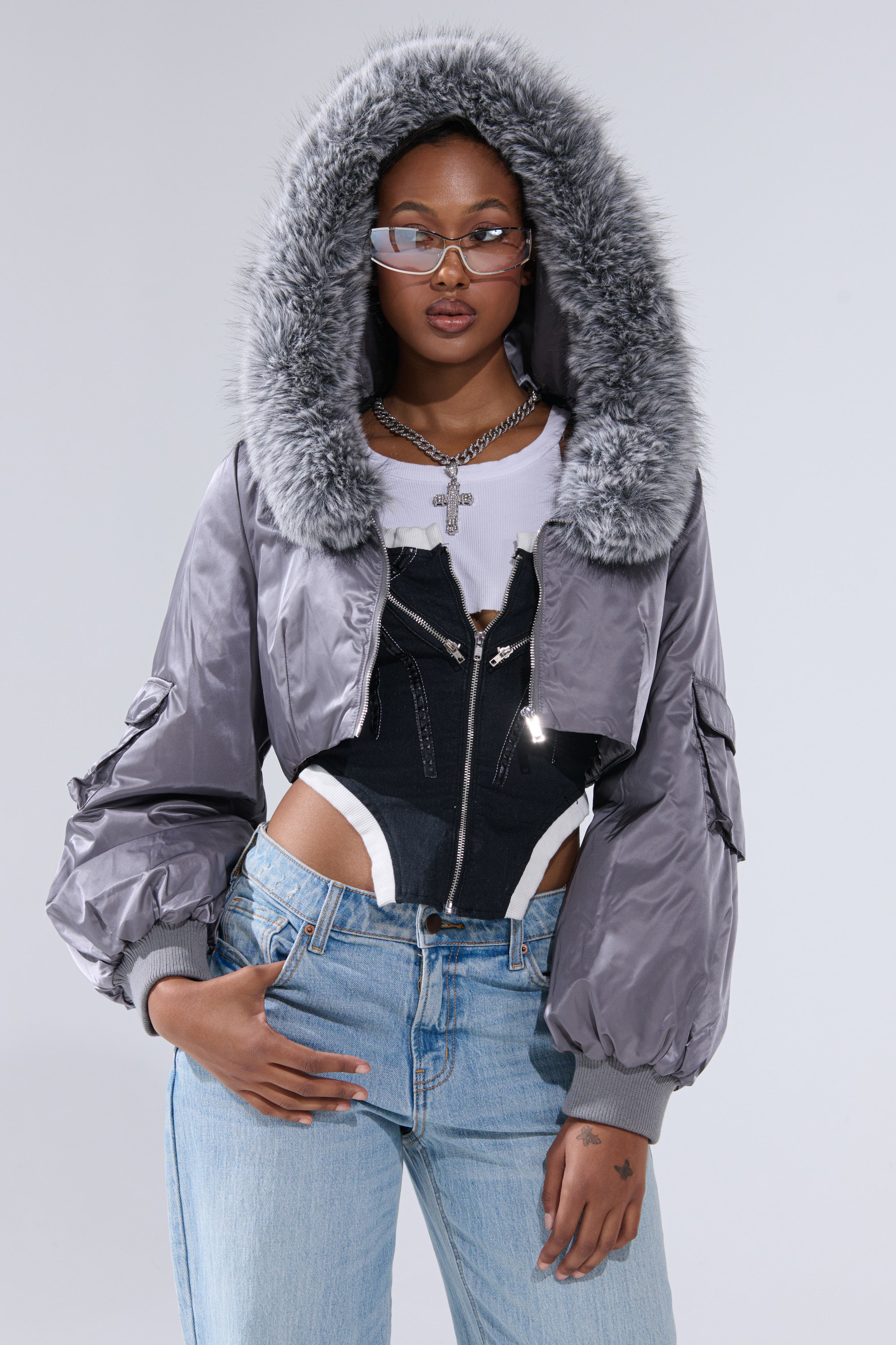 A woman models the ITTY BITTY CROPPED BOMBER in gray with fur trim, paired with a black corset, white top, blue jeans, glasses, and silver jewelry against a plain light backdrop.