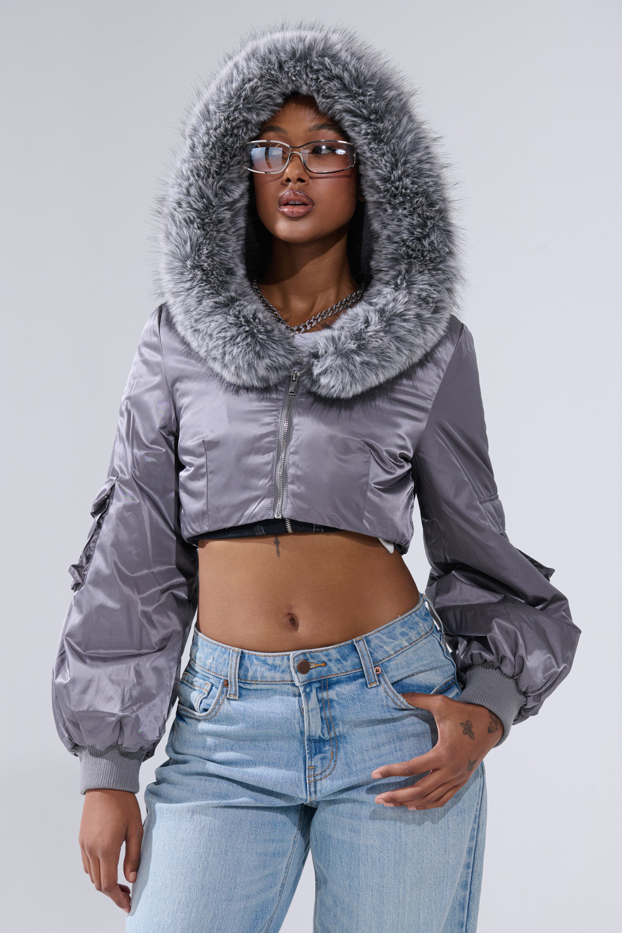 Wearing the ITTY BITTY CROPPED BOMBER—a shiny silver jacket with a large fur trim hood—a person in glasses poses confidently with one hand on their hip, paired with light blue jeans against a plain backdrop.