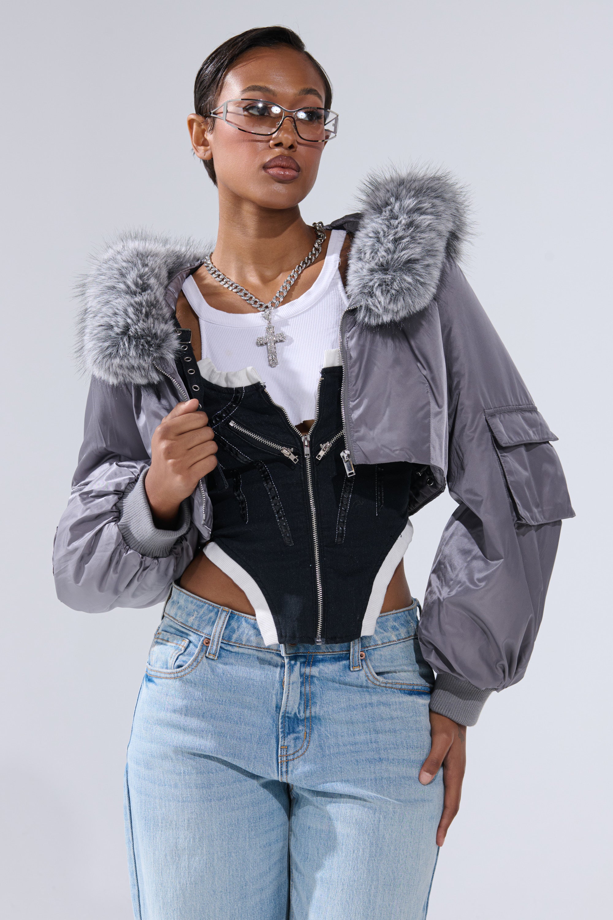 A stylish person wearing glasses, the ITTY BITTY CROPPED BOMBER with fur trim, layered black and white tops, light blue jeans, a silver chain necklace, and a confident look against a plain background.
