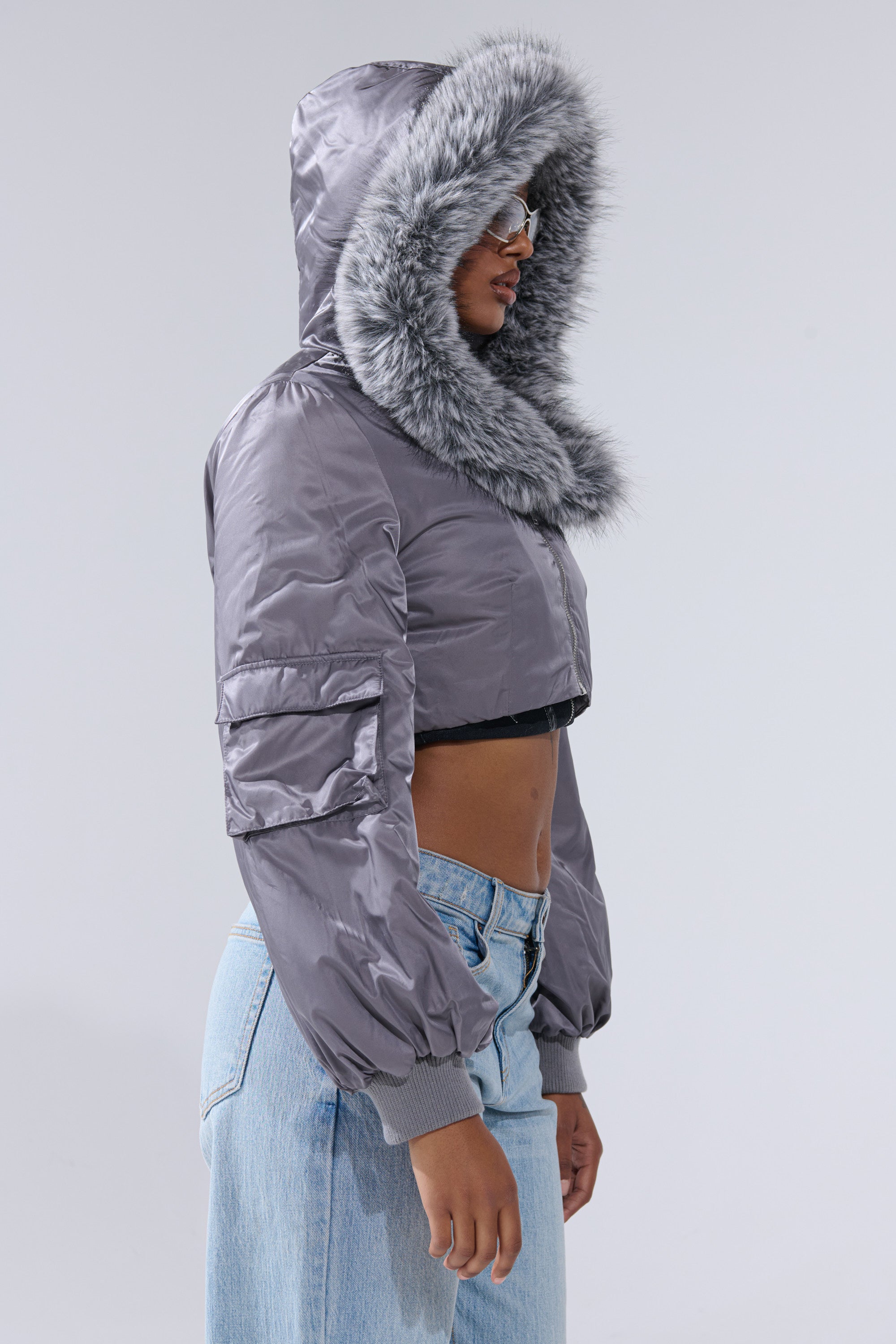 A person stands in profile against a plain light background, wearing the ITTY BITTY CROPPED BOMBER—a shiny gray bomber jacket with fur trim—paired with light blue jeans.