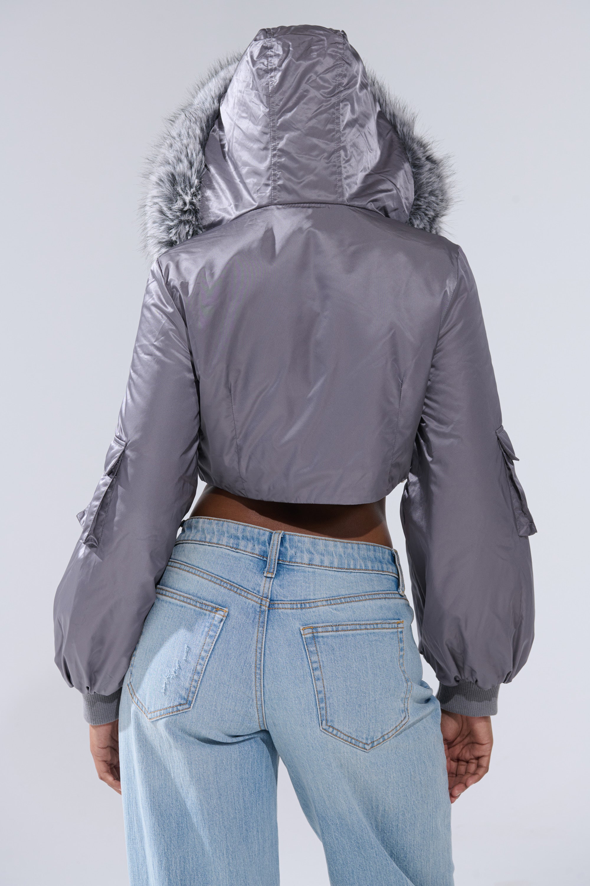 A person wears light blue jeans and the ITTY BITTY CROPPED BOMBER—a shiny silver cropped jacket with a cozy fur-trimmed hood—photographed from the back against a plain light background, showcasing standout street-style outerwear.