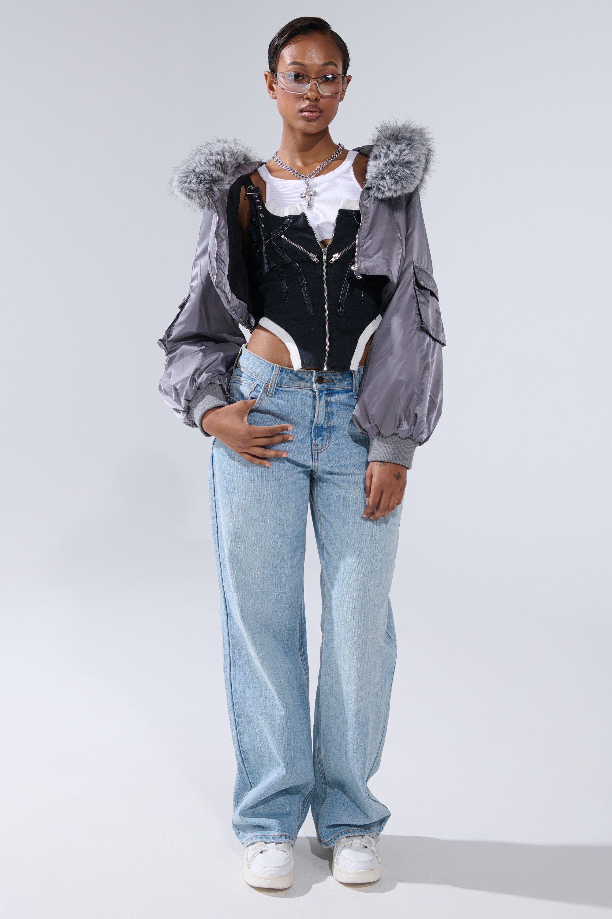 A person stands facing forward, wearing glasses, the ITTY BITTY CROPPED BOMBER with silver fur trim over a white crop top and black corset-style top, light blue wide-leg jeans, and white sneakers, hands resting on their jeans.