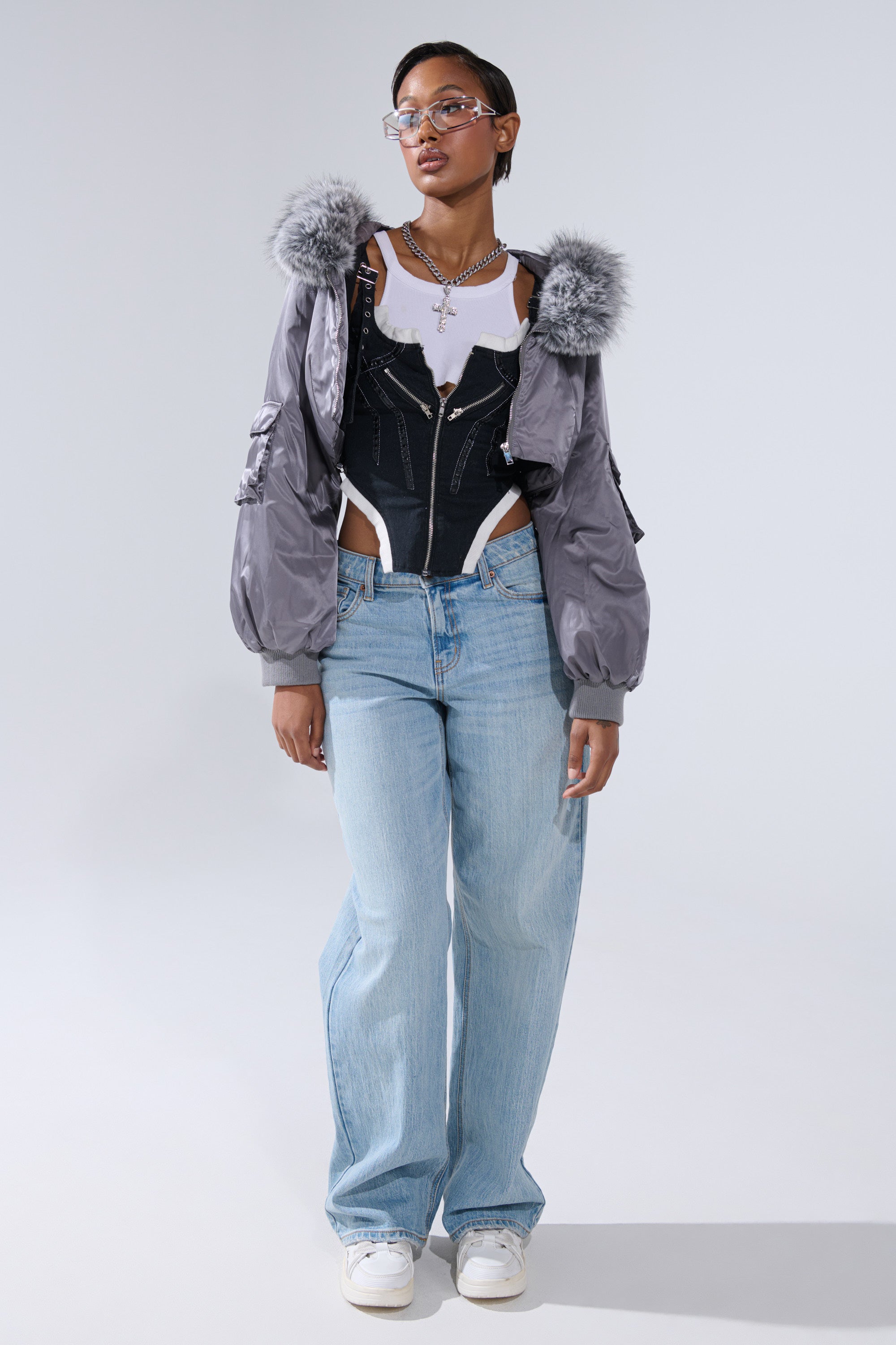 A person wears the ITTY BITTY CROPPED BOMBER—gray with fur-trimmed hood—over a white tee, black corset, and light blue wide-leg jeans, styled with white sneakers, clear glasses, and layered necklaces for a chic street look.