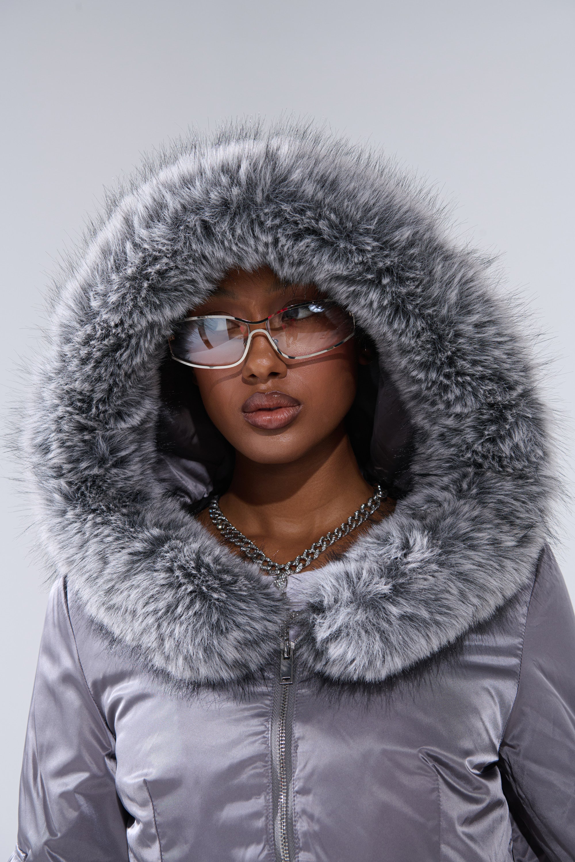 Someone stands against a plain light background wearing the ITTY BITTY CROPPED BOMBER, a shiny gray cropped jacket with thick faux fur trim, paired with translucent glasses and a silver chain necklace.