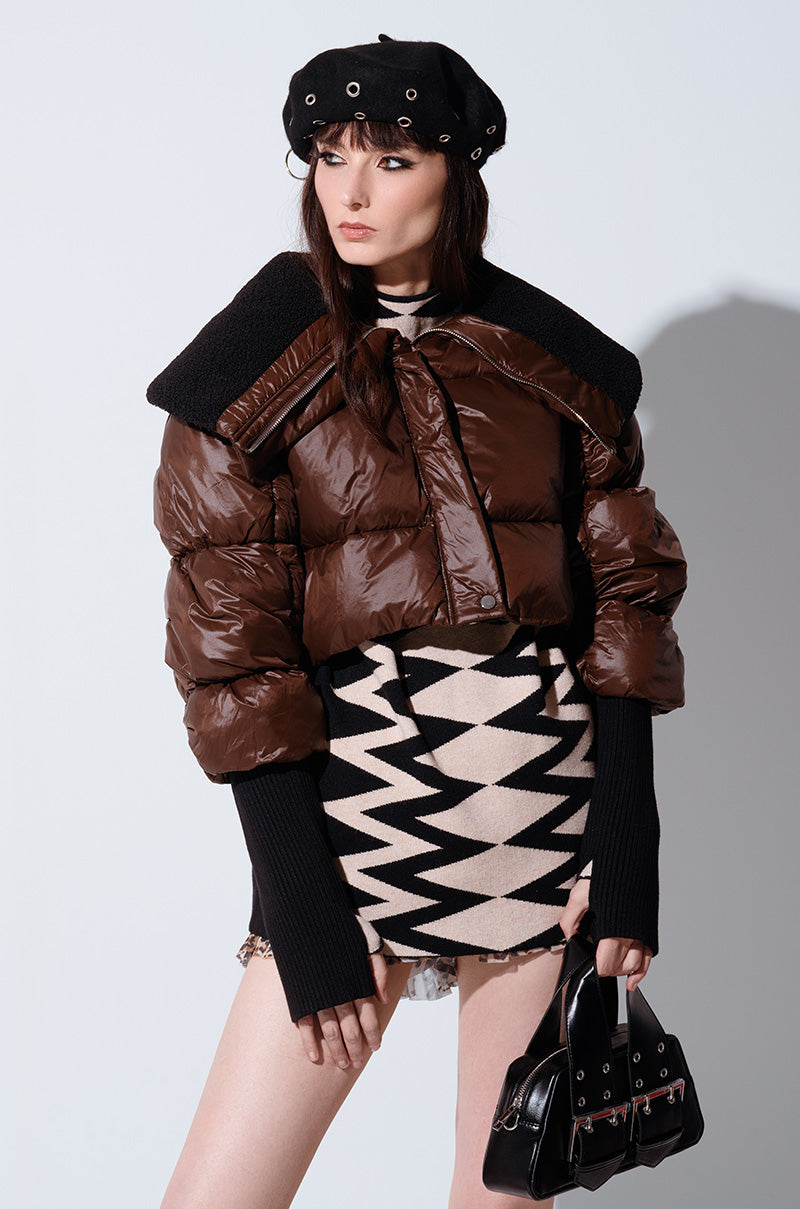 A person poses against a light background wearing the NOLITA CROPPED BOMBER, a glossy brown puffer jacket with a metallic zip-button front closure. They pair it with a patterned black and white dress, accessorize with a black beret and gloves, hold a black handbag, and have long hair with bangs.