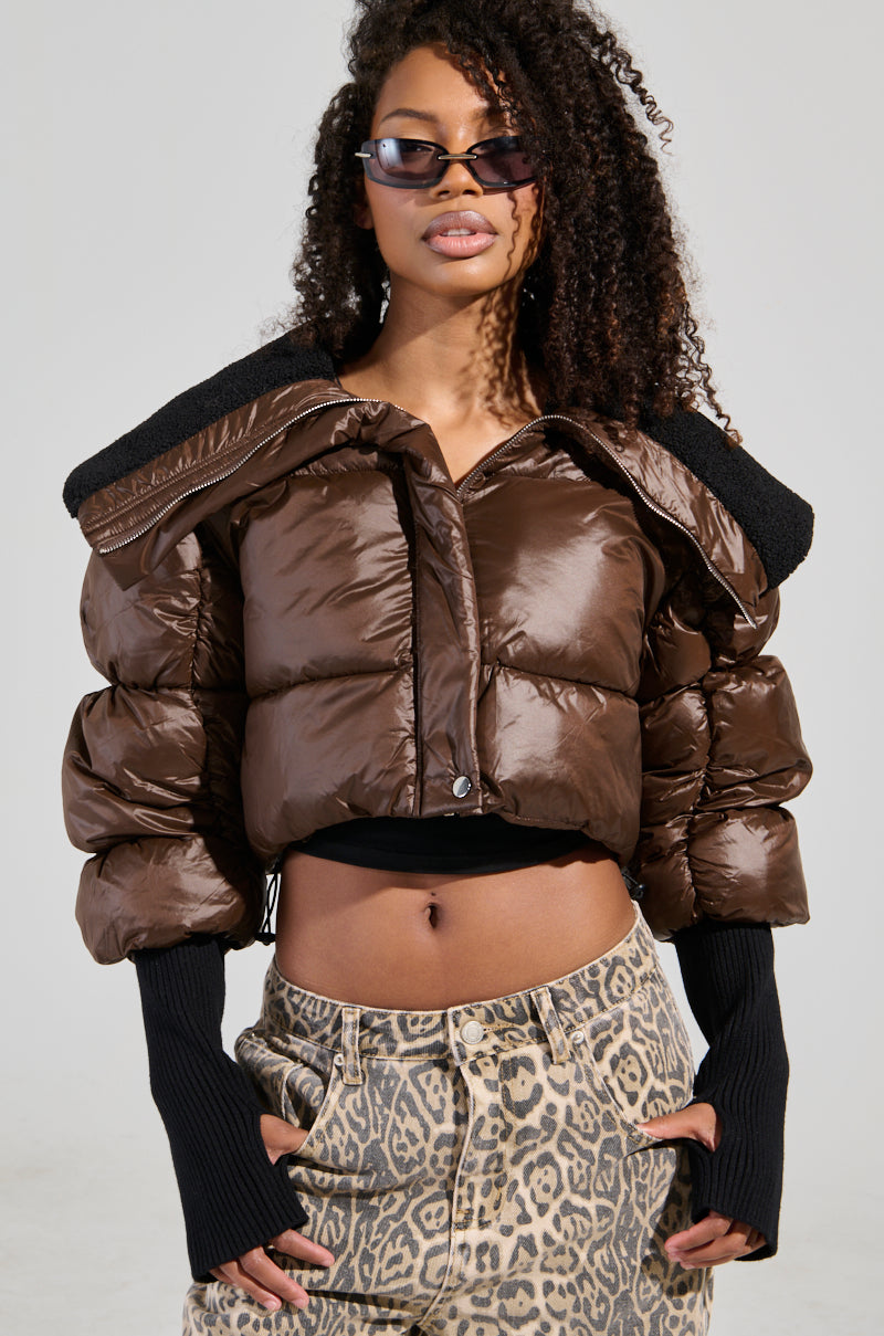 Dressed in a radiant NOLITA CROPPED BOMBER, this woman exudes confidence with its faux sherpa-lined collar and metallic zip-button front closure. She complements her look with black fingerless gloves and leopard print pants, while her curly hair cascades around her sunglasses, creating a striking image against a neutral backdrop.