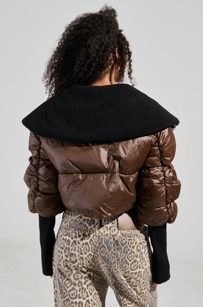 A person with curly hair models the Nolita Cropped Bomber, featuring textured fabric and long sleeves, in a brown puffer style with an oversized black collar, paired with leopard print pants. The individual is viewed from the back.
