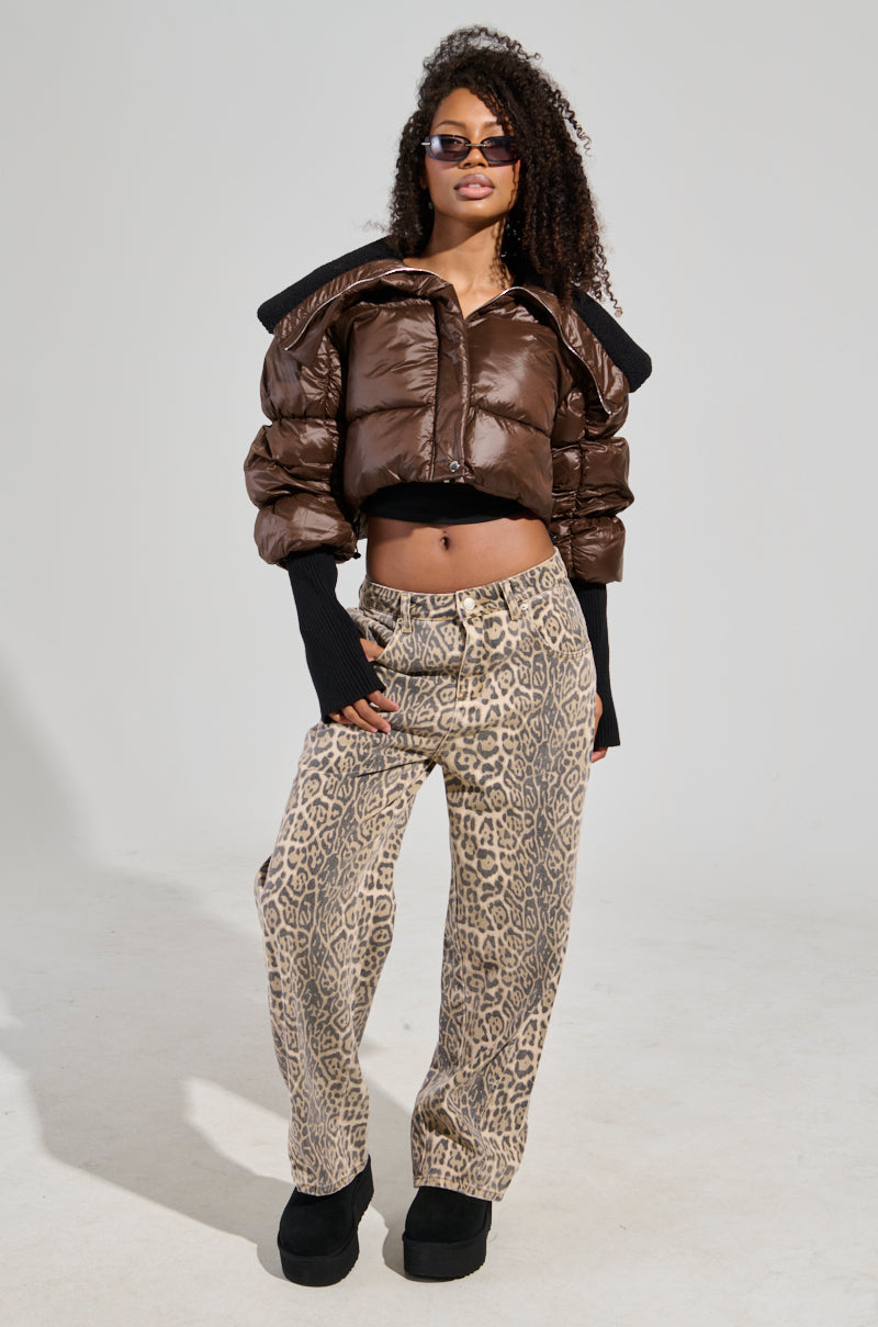 A person with long curly hair and sunglasses is showcasing the NOLITA CROPPED BOMBER, a shiny brown puffer jacket featuring a faux sherpa-lined collar. They pair it with leopard print pants and black platform shoes. The jacket's metallic zip-button front closure introduces an element of chic sophistication against the plain background.
