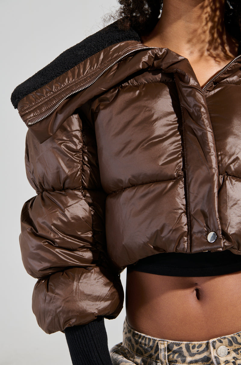A person dressed in a brown NOLITA CROPPED BOMBER with black lining and a faux sherpa-lined collar pairs it with leopard-print pants. The jacket, highlighted by its metallic zip-button front closure, is the focus of the image which showcases the upper half of the outfit against a plain background.