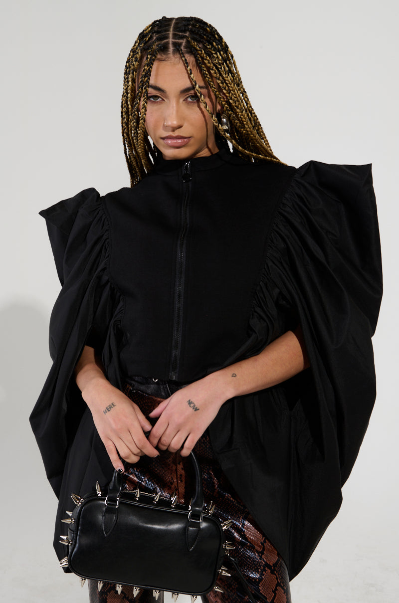 A person with long braided hair wears an oversized black "NOUVEAU RICHE EXAGGERATED SLEEVE BOMBER" paired with a brown patterned skirt. They hold a small, spiked black handbag, resting their tattooed hands gently on it against a plain, light background.