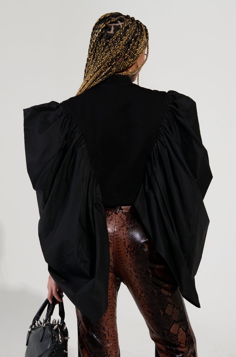 A person with long braids, seen from behind, is dressed in the NOUVEAU RICHE EXAGGERATED SLEEVE BOMBER paired with brown snake-print pants. They carry a black handbag featuring silver details, creating a chic cropped fit that enhances their bold style.