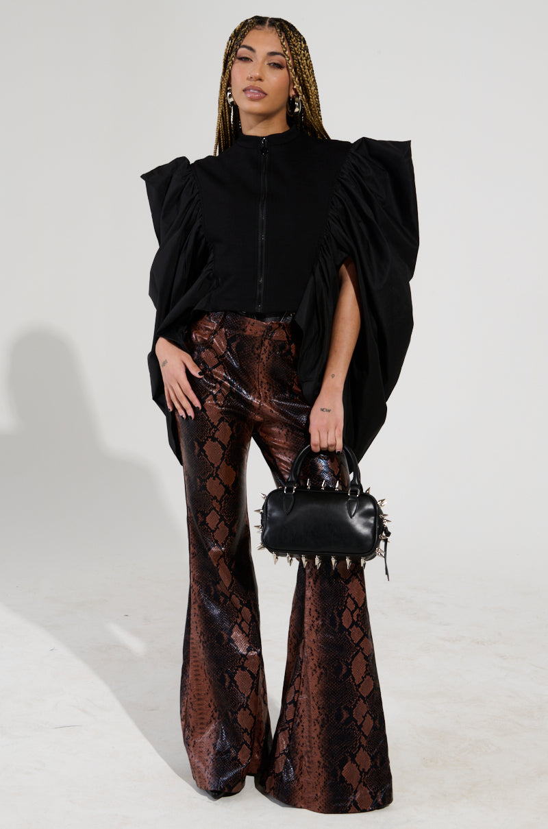 A person with braided hair stands confidently, wearing the NOUVEAU RICHE EXAGGERATED SLEEVE BOMBER in black. They pair it with brown snake-patterned flared pants and hold a small black handbag adorned with spikes against a plain white background.