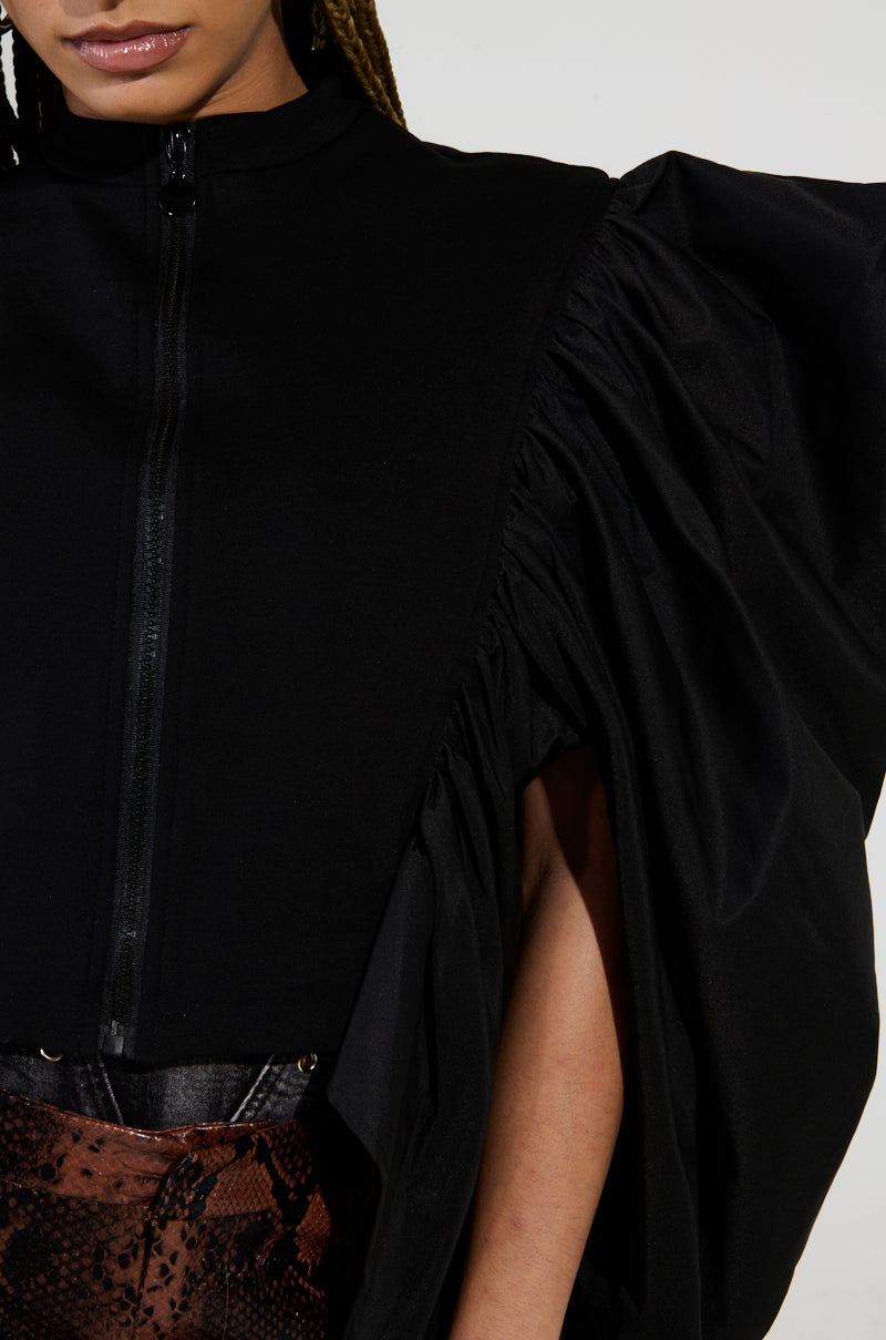 A close-up of an individual wearing the NOUVEAU RICHE EXAGGERATED SLEEVE BOMBER, showcasing its black design with dramatic ruched puff sleeves and a front zipper. It is styled with a cropped fit and high neckline, paired alongside a brown and black snake print skirt. The image is cropped to partially show the face.
