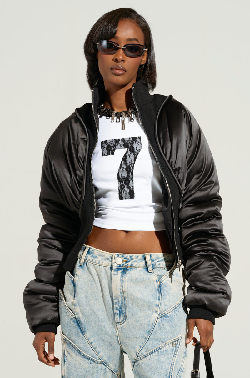A person wearing dark sunglasses, the ELECTRA SATIN EFFECT BOMBER with a cropped silhouette, a white cropped top featuring the number 7, and light wash jeans stands confidently against a plain background. The look is accessorized with a chunky necklace.