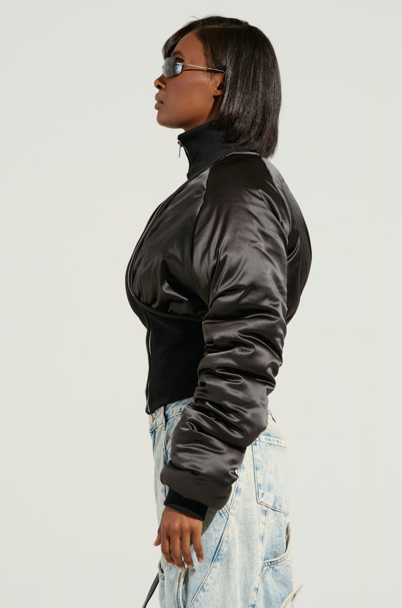 Side profile of a person wearing the ELECTRA SATIN EFFECT BOMBER, paired with light blue jeans and sunglasses. They have short, black hair and are standing against a light gray background. The cropped silhouette of the bomber jacket adds a modern twist to this stylish look.
