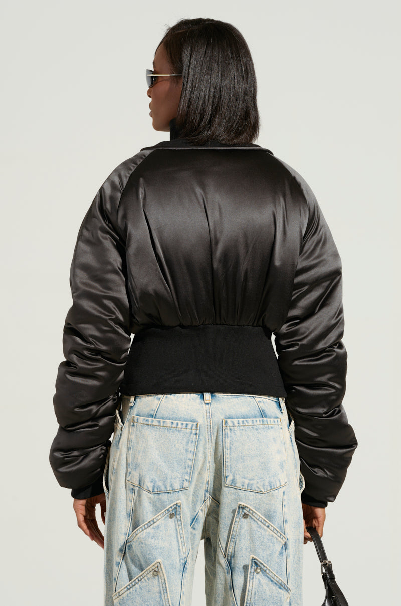 A person wearing the ELECTRA SATIN EFFECT BOMBER jacket and high-waisted, light blue jeans with distinctive pocket stitching stands facing away. They are holding a small black bag and wearing sunglasses, showcasing a chic yet effortless style.
