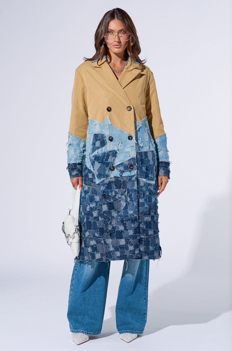 A model stands against a plain background wearing an AKIRA Label Wicker Park Denim Detail Trench with a beige upper half and patchwork blue denim detail lower half. She pairs it with blue denim jeans, white pointed-toe shoes, glasses, and holds a small white handbag.