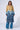 A model stands against a plain background wearing an AKIRA Label Wicker Park Denim Detail Trench with a beige upper half and patchwork blue denim detail lower half. She pairs it with blue denim jeans, white pointed-toe shoes, glasses, and holds a small white handbag.