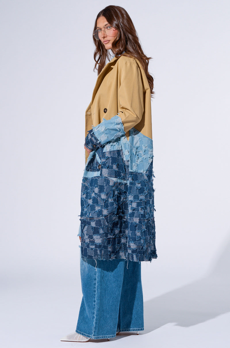A woman with long brown hair wearing glasses stands in an AKIRA Label Wicker Park Denim Detail Trench over wide-leg blue jeans and white shoes. She is turned to her right, gazing forward in a well-lit studio setting with a plain background.