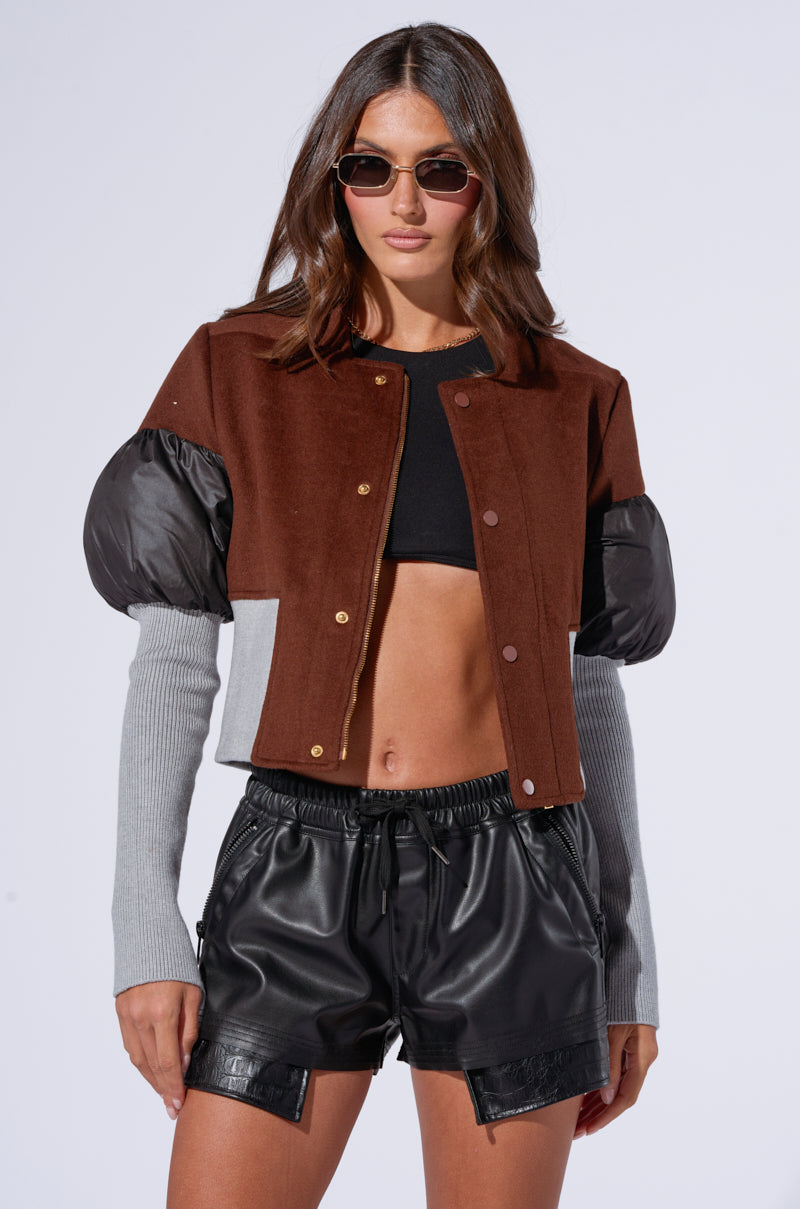 A woman with long brown hair wearing sunglasses poses in a stylish outfit. She dons the OAKLAND MIXED MEDIA BOMBER, featuring a mix of brown and black colors with puffy sleeves, paired with a black cropped top and black leather shorts. The background is a plain white studio backdrop.