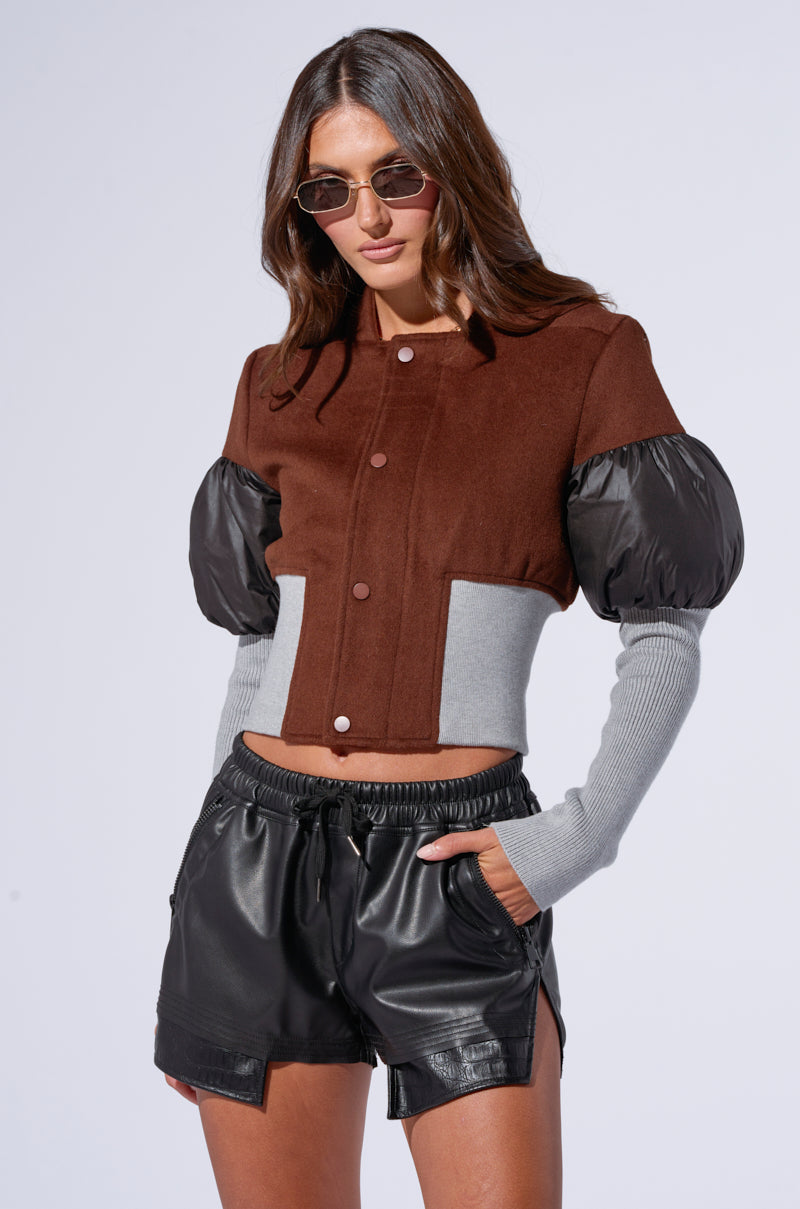 A person with long hair and sunglasses models a stylish outfit featuring the Oakland Mixed Media Bomber, which is a brown and gray wool-blend cropped jacket with puffy black sleeves that exude a bomber style. They pair it with sleek black leather shorts against a plain white background.