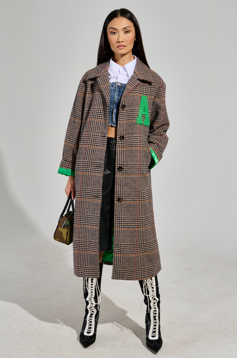 A woman exudes confidence in the TAKE IT EASY RELAXED FIT TRENCH, an oversized coat with green accents made from a wool-blend material. She layers it over a white shirt and denim jacket, complementing the look with a black skirt and white lace-up boots. A small handbag hangs by her side as her long dark hair cascades down against the plain gray background.